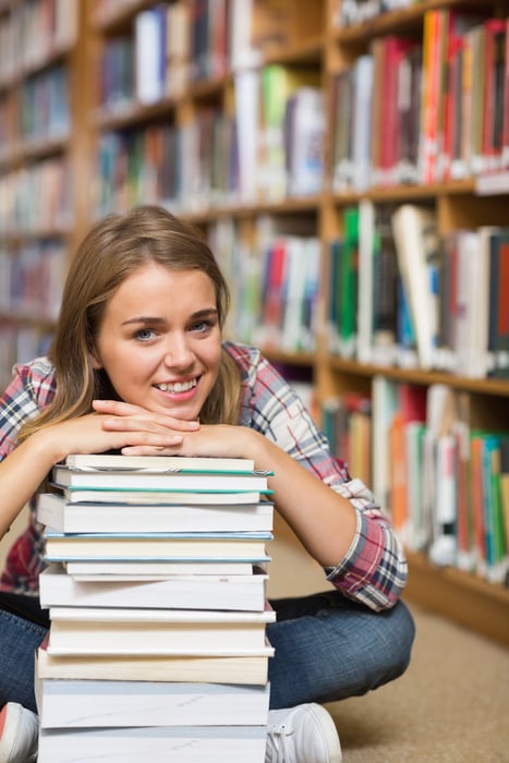 Happy student sitting on library floor leaning on pile of books in college